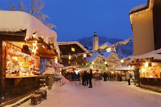 Ein malerischer Weihnachtsmarkt in einem verschneiten Dorf. Verkaufsstände sind festlich dekoriert, und Lichter glitzern in der Dämmerung.