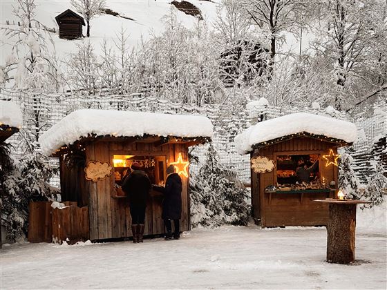 Ein winterlicher Weihnachtsmarkt mit zwei Holzbuden, die geschmückt und beleuchtet sind. Der Boden ist mit Schnee bedeckt, und im Hintergrund sind verschneite Bäume zu sehen.