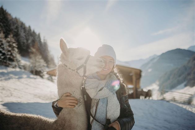 Eine Frau umarmt ein Lama in einer schneebedeckten Landschaft. Im Hintergrund sind Berge und ein Holzhaus sichtbar.