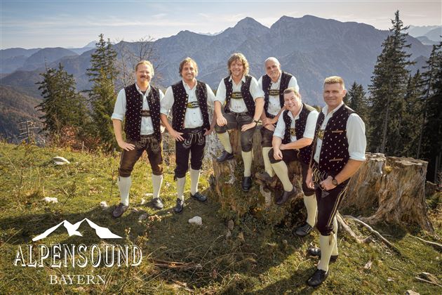 Eine Gruppe von sechs Männern in traditioneller bayerischer Tracht steht auf einem Berg. Im Hintergrund sind beeindruckende Alpenlandschaften zu sehen.