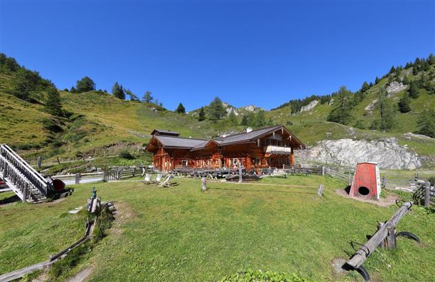 Eine gemütliche Berghütte umgeben von grünen Wiesen und hohen Bergen. Der Himmel ist klar und blau.
