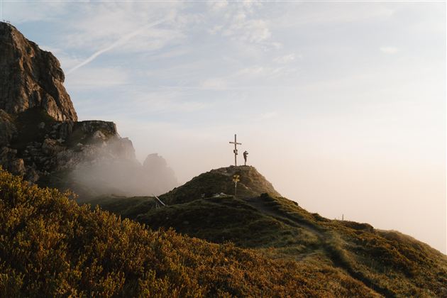 Eine neblige Landschaft mit einem Kreuz auf einem Hügel. Im Hintergrund sind Felsen und der Himmel sichtbar.