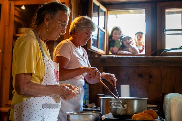 Zwei Frauen kochen in einer gemütlichen Küche. Im Hintergrund schauen einige Kinder neugierig durch das Fenster.