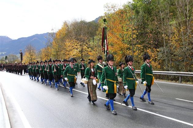 Eine Parade mit uniformierten Teilnehmern, die in bunten Trachten marschieren. Im Hintergrund sind herbstliche Bäume und Berge zu sehen.