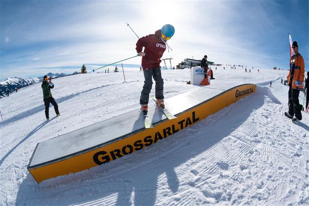 Eine Person fährt auf Skiern über ein Snowboard-Rail in einer verschneiten Berglandschaft. Im Hintergrund sind weitere Skifahrer und eine wunderschöne alpine Kulisse zu sehen.