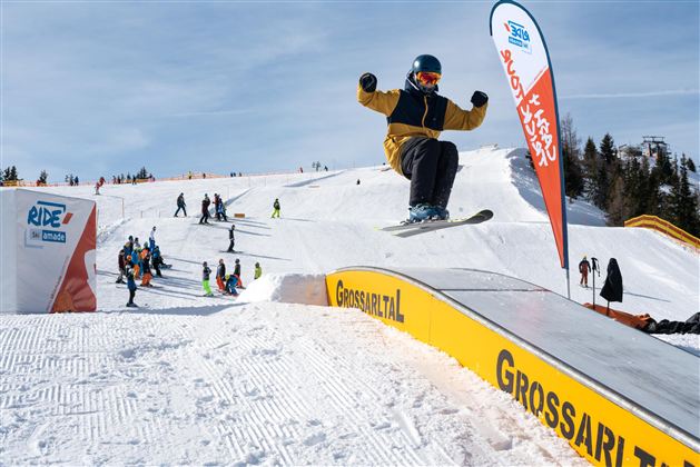 Ein Snowboarder springt über eine Rampe im Skigebiet Grossarltal. Im Hintergrund sind weitere Skifahrer und Skilehrer sichtbar.