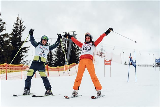 Zwei Skifahrer freuen sich mit erhobenen Händen auf einer verschneiten Piste. Im Hintergrund sind Bäume und Tore zu sehen.