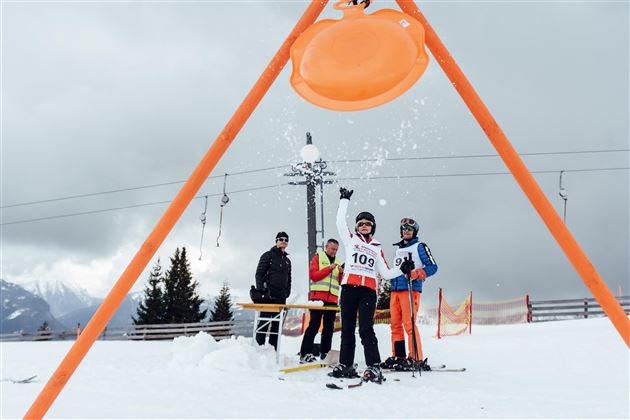 Ein Junge steht auf Skiern und jubelt, während Schnee herunterfällt. Im Hintergrund sind zwei weitere Personen und eine Skiliftanlage zu sehen.
