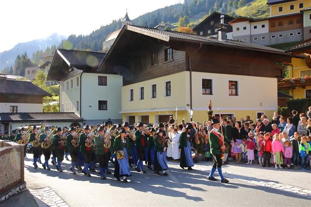 Ein Festumzug mit Musikkapellen marching durch eine malerische Straße. Im Hintergrund sind traditionelle Häuser und viele Zuschauer, darunter Kinder, zu sehen.