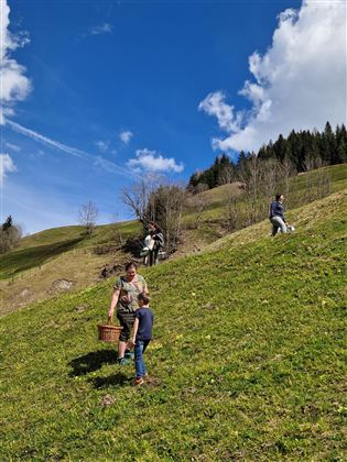A green meadow with some trees and a blue sky. A person is carrying a basket, while a child stands nearby.