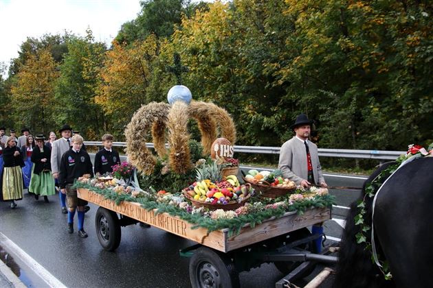 Eine festliche Parade mit einem Wagen, der mit Obst und Erntegaben geschmückt ist. Menschen in traditionellen Trachten begleiten den Wagen entlang der Straße.