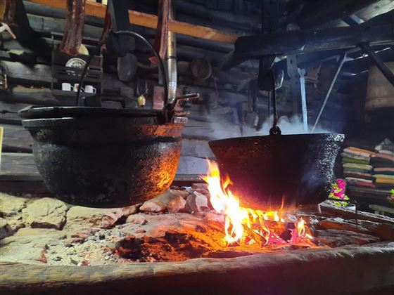 Ein rustikaler Holzofen mit zwei hängenden Kochtöpfen über einem offenen Feuer. Die Flammen leuchten warm und schaffen eine gemütliche Atmosphäre.