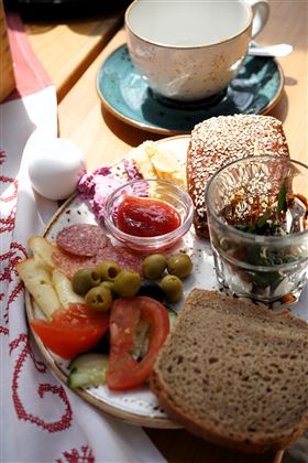 Ein gedeckter Tisch mit einem Frühstücksplateau, das Brot, Wurst, Gemüse und ein Glas mit Kräutern enthält. Daneben steht eine Tasse und ein Stück Obst.