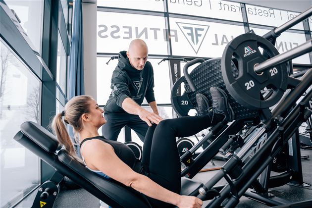 Eine Frau trainiert im Fitnessstudio, während sie Unterstützung von einem Trainer erhält. Die Geräte und die große Fensterfront sorgen für ein modernes Ambiente.