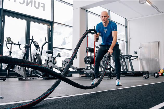 Ein Mann trainiert im Fitnessstudio mit einem Battle Rope. Der Raum ist modern und gut ausgestattet mit verschiedenen Fitnessgeräten.