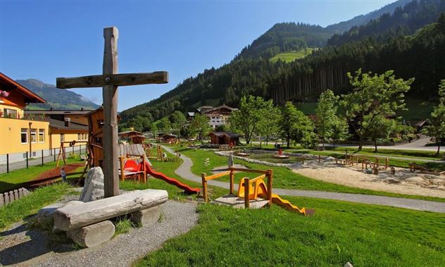 Ein schöner Spielplatz in einer grünen Umgebung mit Bergen im Hintergrund. Es gibt Rutschen, Schaukeln und einen holzernen Kreuz als Blickfang.