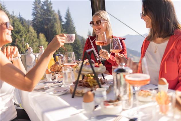 Three women enjoy breakfast outdoors with a view of the mountains. They toast with glasses and laugh together.