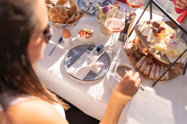 A nice breakfast with various foods and drinks on a table. In the foreground, there is a person sitting at the table and eating enjoyably.