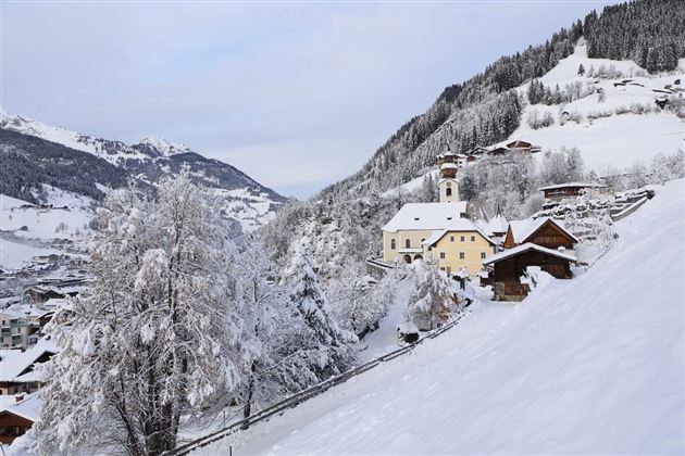 Eine verschneite Landschaft mit einem charmanten Dorf und einer Kirche. Im Hintergrund erstrecken sich die Berge unter einem grauen Himmel.