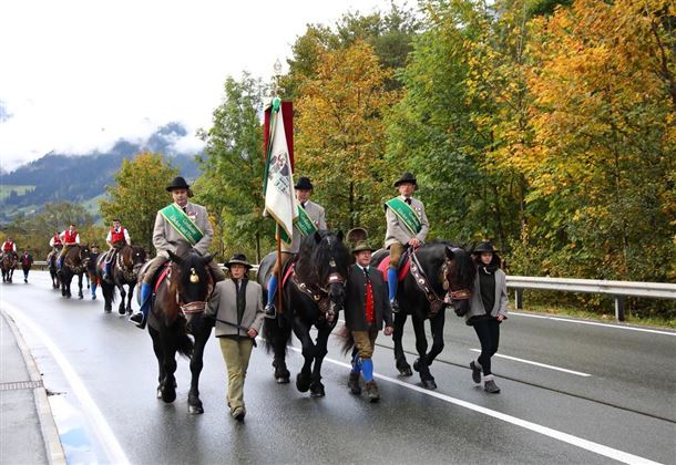 Eine Gruppe von Reitern in traditioneller Kleidung zieht auf einer Straße entlang. Die Umgebung zeigt bunte Herbstbäume und eine leicht bewölkte Himmel.