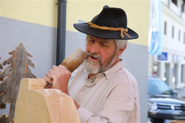 An older man with a hat is carving wood. He is working on a large piece of wood in an urban setting.
