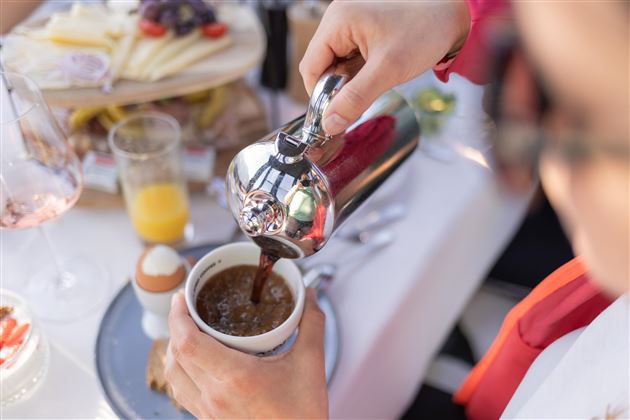 A person is pouring freshly brewed coffee into a cup. In the background, some breakfast treats and drinks can be seen.