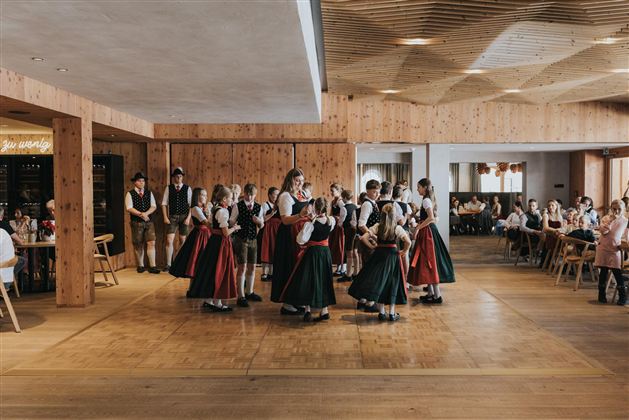 Eine Gruppe von Tänzern in traditionellen Trachten tanzt in einem hellen Saal. Im Hintergrund sitzen Gäste an Tischen und beobachten die Darbietung.