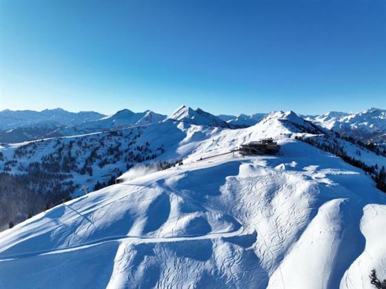 Eine ruhige Winterlandschaft mit schneebedeckten Bergen unter einem klaren blauen Himmel. Skispuren ziehen sich über die weiße Oberfläche.