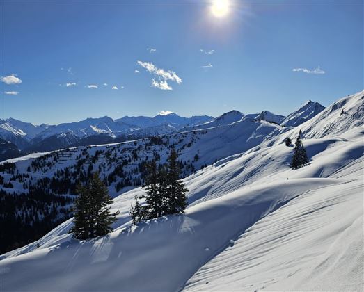 Eine schneebedeckte Berglandschaft unter blauem Himmel. Die Sonne scheint hell und die Berge erstrecken sich im Hintergrund.