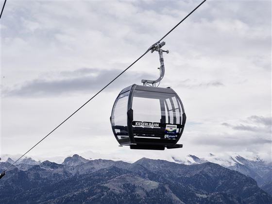 Eine moderne Gondelbahn schwebt über die Berge. Der Himmel ist bewölkt und die Berglandschaft ist majestätisch.