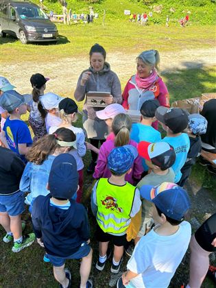 A group of children listens attentively to two women who are explaining something to them. In the background, trees and a meadow can be seen.