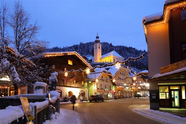 Eine malerische Winterszene mit schneebedeckten Häusern und festlicher Beleuchtung. Im Hintergrund steht eine Kirche auf einem Hügel, umgeben von Bergen.