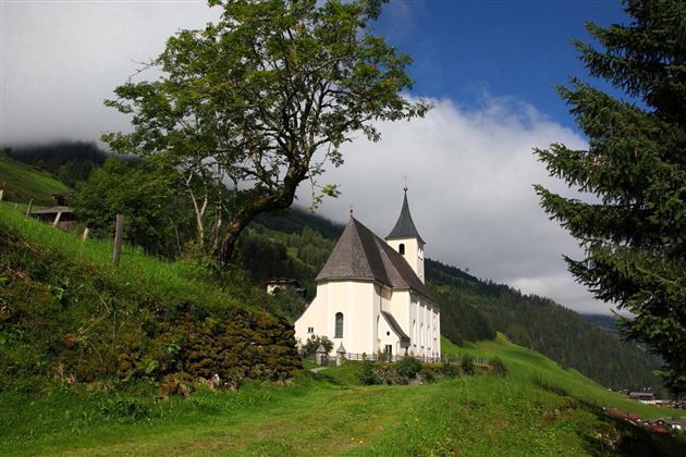Eine malerische Kirche umgeben von üppigem Grün und sanften Hügeln. Im Hintergrund sind bewaldete Berge und ein klarer Himmel zu sehen.