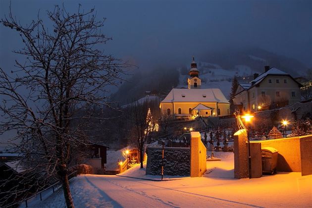 Eine verschneite Landschaft bei Nacht mit einer Kirche und beleuchteten Häusern. Der Himmel ist bewölkt und die Lichter erzeugen eine warme Atmosphäre.
