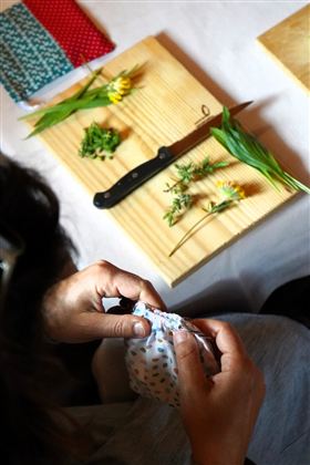 Eine Person hält einen bunten Stoffbeutel in den Händen und näht daran. Auf einem Tisch liegen frische Kräuter und Blumen neben einem Messer und einigen Stoffstücken.