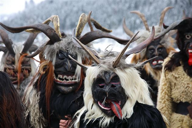 Ein traditionelles Fest mit maskierten Personen in auffälligen Kostümen und Hörnern. Der Hintergrund zeigt eine verschneite Landschaft.
