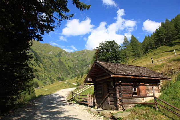 Eine gemütliche Hütte aus Holz liegt am Rand eines Schotterweges. Im Hintergrund erstrecken sich grüne Berge unter einem klaren blauen Himmel.
