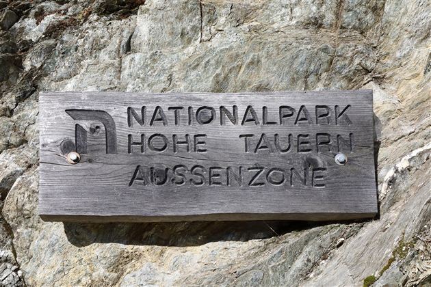 Ein Holzschild mit der Aufschrift "NATIONALPARK HOHE TAUERN AUSSENZONE". Es befindet sich an einem felsigen Hintergrund.