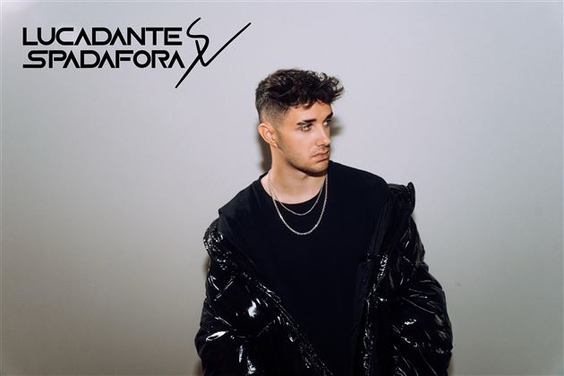 Ein junger Mann mit kurzen, gewellten Haaren trägt eine schwarze Jacke und Ketten. Der Hintergrund ist neutral und hebt seine Erscheinung hervor.