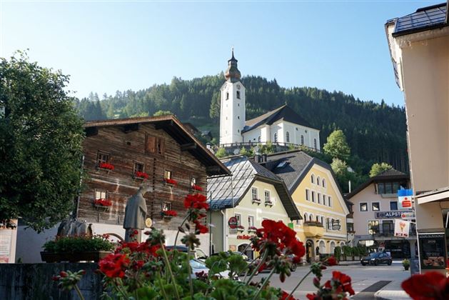 Eine malerische Dorfansicht mit traditionellen Chalets und einer Kirche auf einem Hügel. Im Vordergrund blühen rote Geranien.