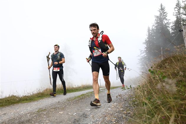 Drei Läufer bewegen sich auf einem schmalen Weg durch neblige Landschaft. Im Hintergrund sind einige Bäume zu sehen.