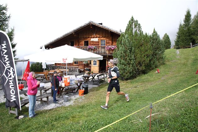 Ein Läufer passiert eine Berghütte, während Zuschauer ihn anfeuern. Im Hintergrund sind Bäume und eine Zeltterrasse zu sehen.