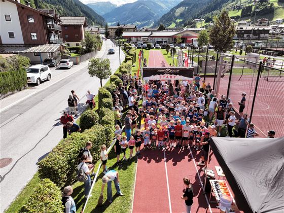 Eine große Gruppe von Kindern steht an der Startlinie eines Laufwettbewerbs. Im Hintergrund sind Berge und eine idyllische Landschaft zu sehen.