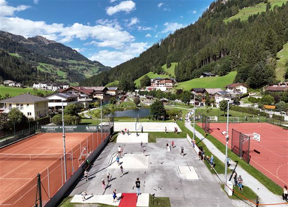 Ein malerischer Blick auf ein Sportgelände mit Tennisplätzen und einem Freizeitraum. Im Hintergrund erstrecken sich grüne Hügel und ein malerisches Dorf.