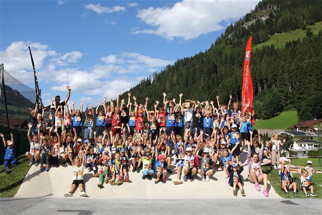 Eine große Gruppe von Menschen steht fröhlich zusammen und hebt die Hände in die Luft. Im Hintergrund sind Berge und eine grüne Landschaft zu sehen.