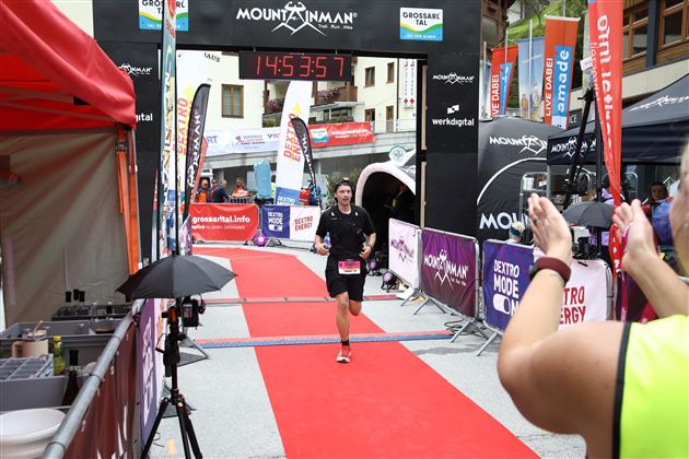 Ein Läufer erreicht das Ziel eines Wettkampfs auf einem roten Teppich. Im Hintergrund sind Banner und jubelnde Zuschauer zu sehen.