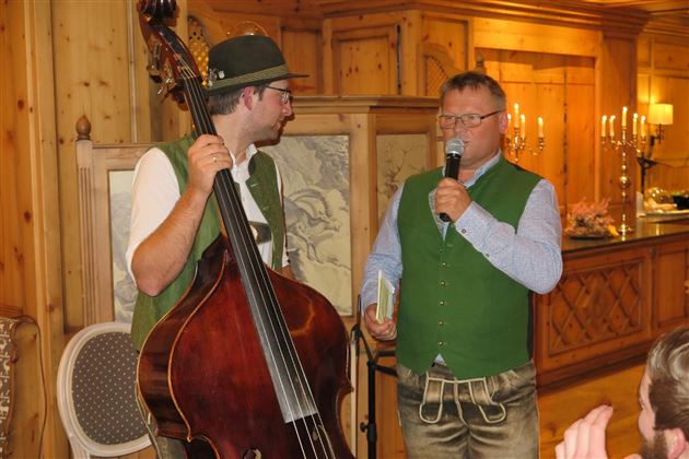Zwei Männer in traditioneller Tracht stehen in einem gemütlichen Raum. Einer spielt Kontrabass, während der andere ein Mikrofon hält und spricht.
