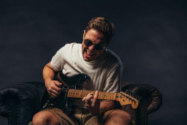 Ein junger Mann spielt begeistert E-Gitarre auf einem Sofa. Er trägt Sonnenbrillen und hat Spaß beim Musizieren.