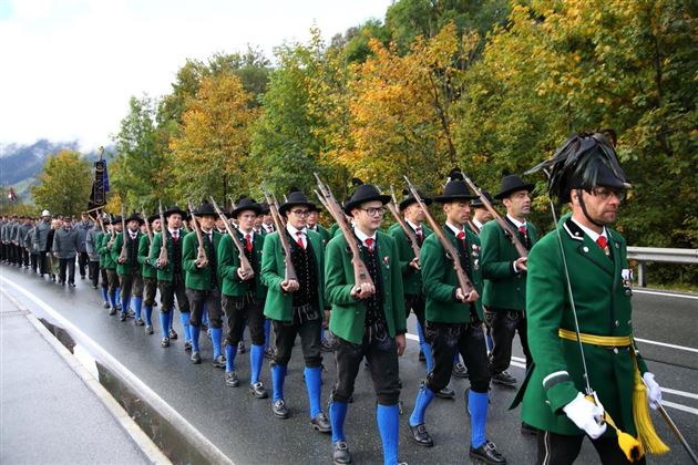 Eine Gruppe von Menschen in traditionellen Trachten marschiert auf einer Straße. Sie tragen Gewehre und sind von bunten Bäumen umgeben.