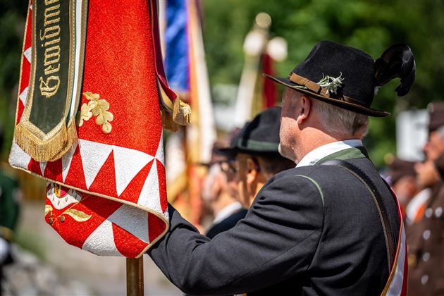 Eine Gruppe von Menschen in traditioneller Kleidung steht und hält Fahnen. Im Hintergrund sind weitere Personen und grüne Bäume zu sehen.